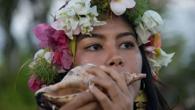 Una mujer de Guam, con una corona de flores tropicales sopla la concha de un caracol marino