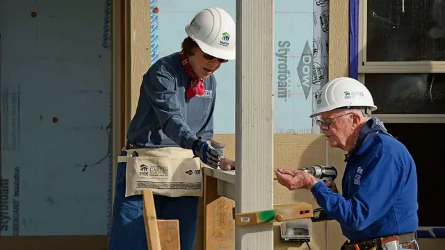 El matrimonio Carter, ambos ya de avanzada edad y vestidos con ropa de trabajo, herramientas y casco, en una construcción. 