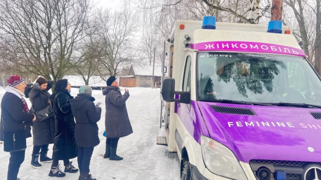 Cinco mujeres abrigadas con gruesos abrigos y gorros permanecen de pie en la nieve frente a la clínica móvil.