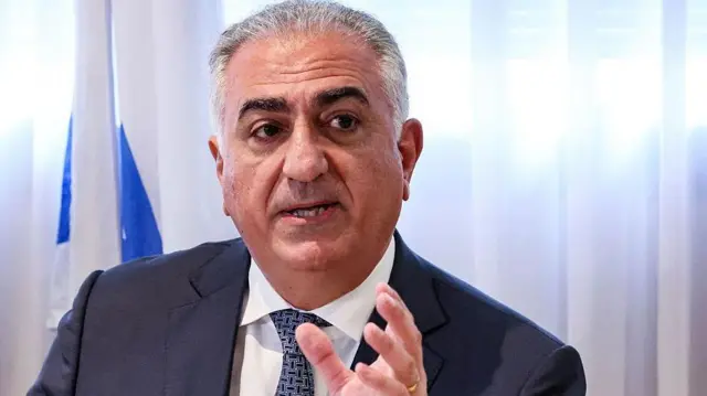 Una imagen de Reza Pahlavi vestido con un traje y cabello canoso haciendo un gesto con una mano levantada.