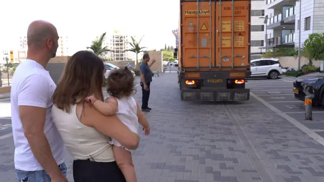 Un camión naranja retrocede mientras Nofar, con su niña en brazos, y Eyal lo observan aproximarse.