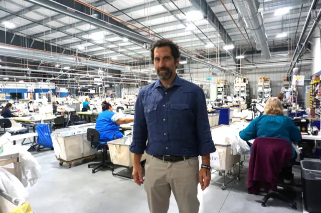 George Matouk, con una camisa azul de manga larga, en su fábrica en Fall River. Detrás de él, unas mujeres están sentadas en sus puestos de trabajo en el amplio almacén.