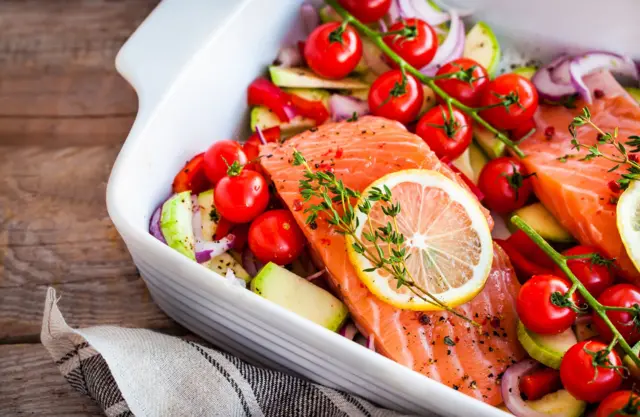 Un salmón en una bandeja con tomates, hierbas y otras verduras, antes de ser horneado