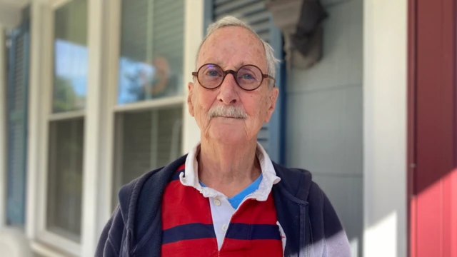 Jeff Stein, con anteojos y bigote blanco, viste un polo a rayas y mira a la cámara delante de una casa