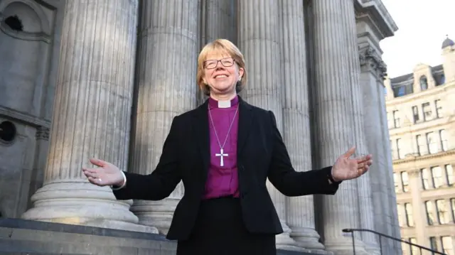 Sarah Mullally con camisa violeta, sonrie abrindo los brazos en frente de la escalinata de la catedral de St. Paul en Londres