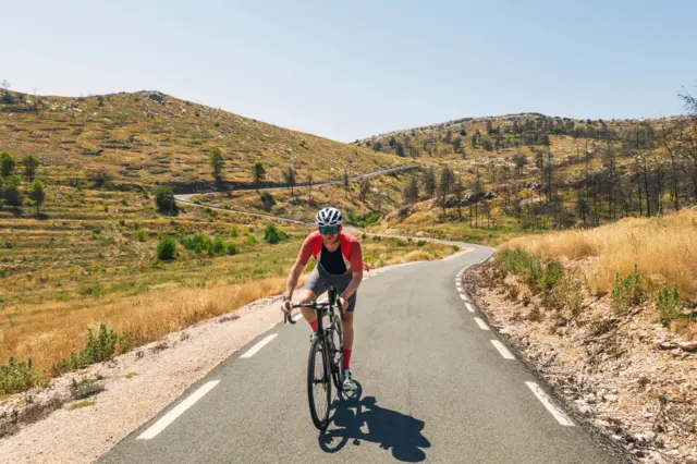 Una persona sobre una bicicleta en un soleado día de verano desciende una serpenteante carretera que baja de dos altos montes. 