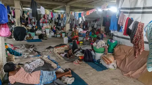 Mujeres y niños en un galpón en medio de colchones en el suelo y ropas tendidas