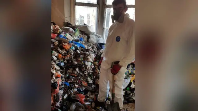 La fotografía muestra una pila de basura apilada en el ventanal. Un joven está de pie frente a la pila. Lleva equipo de protección personal completo, mascarilla y una pala en la mano. Lleva guantes negros y rojos.