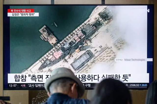 En una fotografía tomada en Seúl el 22 de mayo de 2025, la gente se sienta cerca de un televisor que muestra imágenes de noticias con una imagen satelital cortesía de Maxar Technologies que muestra una vista general de un nuevo buque de guerra norcoreano en el puerto antes de su botadura en Chongjin.