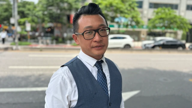 Stanley Wei, vestido con camisa blanca, corbata y chaleco azules, con gafas y el pelo oscuro peinado hacia arriba.