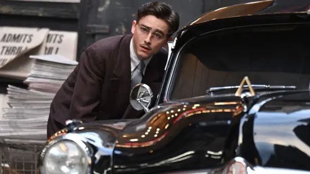 Timothée Chalamet vestido con traje y gafas e inclinándose hacia la ventanilla de un taxi.