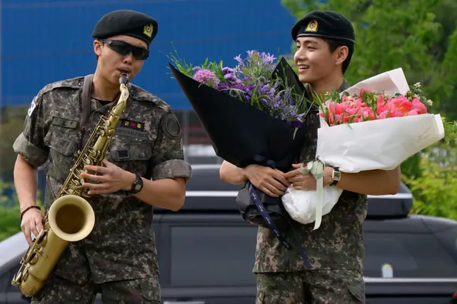 Los miembros de la banda de K-pop BTS, RM (izquierda) toca el saxofón mientras V observa sosteniendo ramos de flores en unas instalaciones deportivas al aire libre en Chuncheon el 10 de junio de 2025.