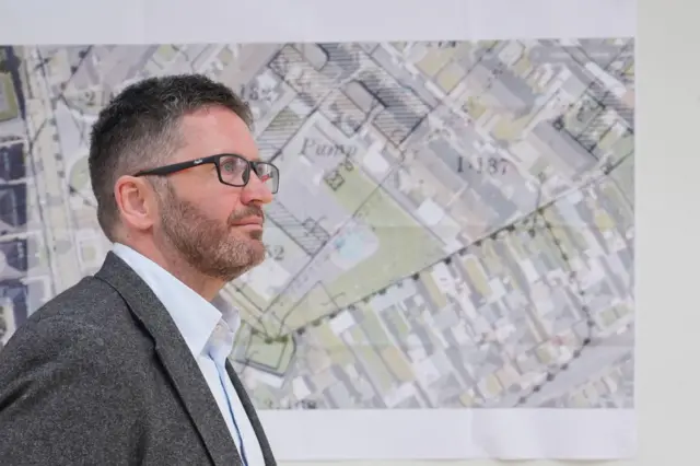 Un hombre con chaqueta gris, camisa clara y gafas mira hacia un lado. Tiene el pelo corto y oscuro con canas. Está de pie frente a un mapa topográfico superpuesto a la imagen de un yacimiento.