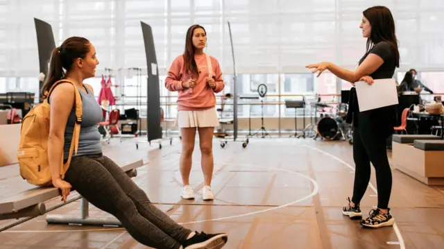 Sammi está de pie en una gran sala de ensayos, ensayando junto a otros dos compañeros en Estados Unidos. Lleva ropa deportiva negra y sus compañeros también llevan ropa deportiva. Al fondo se ve a gente de pie junto a un piano en el que toca un hombre.