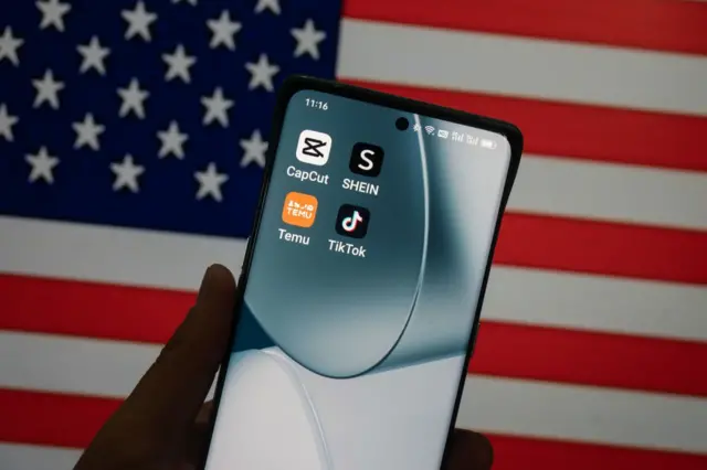 Una mano sostiene un teléfono donde se ven los logos de Temu y Shein y en el fondo la bandera de EE.UU.