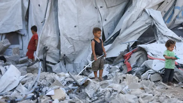 Dos niños y una niña, en pie sobre una montaña de escombros junto a tiendas de campañas en un asentamiento de desplazados en Ciudad de Gaza. La niña tiene en las manos un juguete.