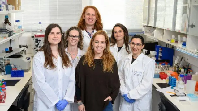 Grupo de seis científicos -cinco mujeres y un hombre- tomándose una foto grupal, la mayoría con batas blancas en el laboratorio del Instituto de Ciencias Weizmann