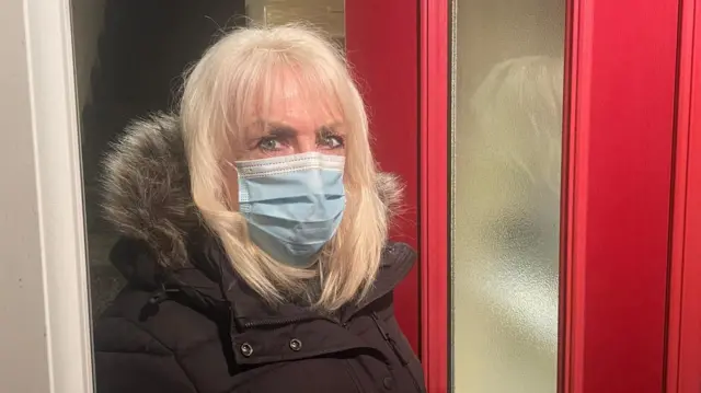 Andrea está parada frente a la puerta de entrada abierta. La puerta es roja y se puede ver una escalera detrás de ella. Lleva un abrigo negro con capucha peluda y una mascarilla azul.