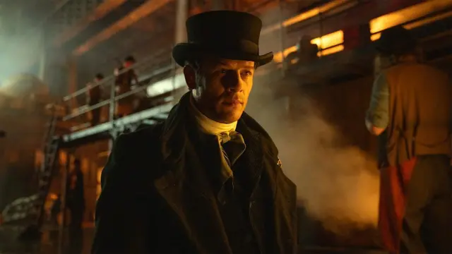 James Norton, vestido con ropa y accesorios de época para darle vida a un personaje, mira detenidamente un punto fuera del cuadro con expresión seria mientras en el fondo transcurre la actividad de una fábrica, como lo sugieren el humo y los que parecen ser trabajadores de la misma.
