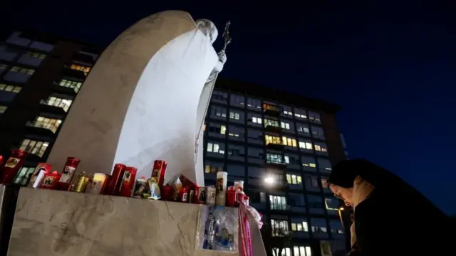 Una monja inlcina la cabeza a los pies de una estatua de mármol del papa cuya base ha sido cubierta de velas, en una imagen nocturna.