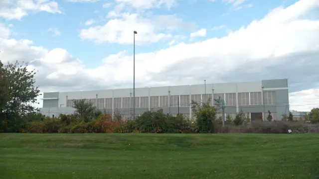 Un gran edificio tipo almacén industrial se encuentra detrás de un campo plano de césped y árboles.