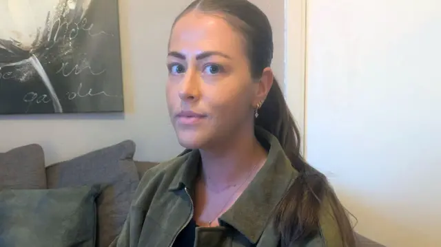 Nadia sentada en un sofá, con chaqueta verde y cabello recogido, junto a un cuadro decorativo en la pared.