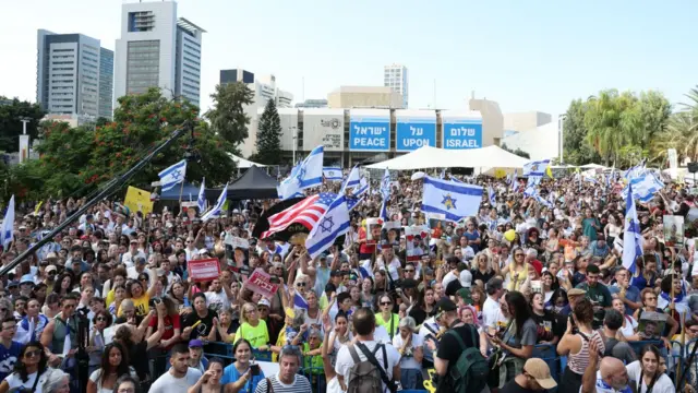 Miles de personas reunidas en un gran espacio abierto, agitando banderas de Israel y una de Estados Unidos. Al fondo, unos carteles piden