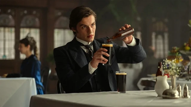 Un hombre joven vestido de época en un elegante comedor sirve una cerveza oscura en un vaso. Su expresión es seria y concentrada.