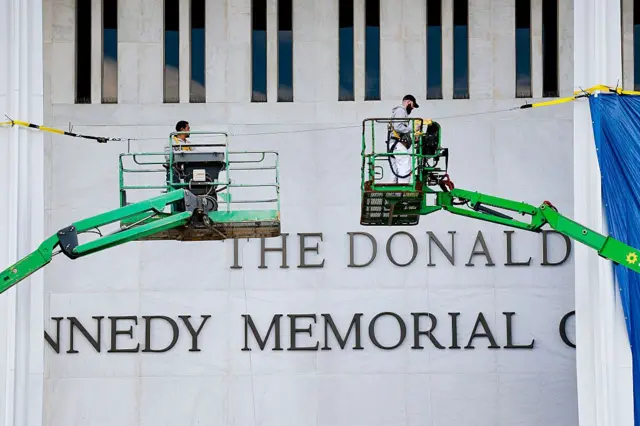 Dos operarios subidos a máquinas para trabajar frente a la fachada del Centro Kennedy, que muestra el nombre de Trump encima del de Kennedy. 