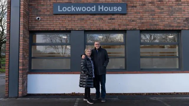Fotografía de Rob y Dianne Parsons frente al Lockwood Centre. Ronnie está a la derecha de Dianne y viste pantalones, gafas y un abrigo negro tipo pea coat. Dianne lleva un abrigo negro acolchado y leggings negros, y sonríe a la cámara. Sobre una fila de ventanas del edificio de ladrillo se puede ver un cartel que dice ‘Lockwood House’.