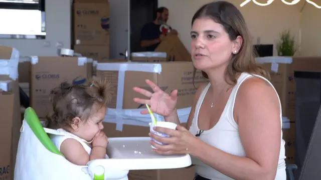 Una mujer rodeada de cajas de cartón mueve las manos mientras parece hablar con alguien. Sujeta la comida que le está dando al bebé sentado frente a ella en una trona.