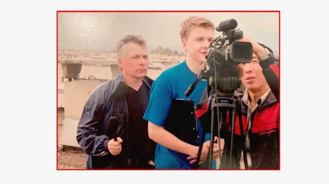 Un equipo de filmación de tres personas, entre ellas el joven Pavel Zarubin, se encuentra junto a una cámara de video. Zarubin lleva una camiseta azul y mira por el visor. Un hombre con una chaqueta roja toca la parte superior de la cámara. Están al aire libre con el horizonte de la ciudad a sus espaldas.
