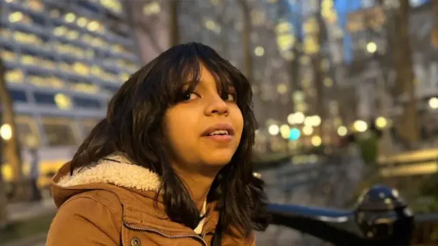 Juliana Peralta, una chica de pelo oscuro, aparece en una fotografía nocturna con una chaqueta marrón con capucha forrada de borrego, con un paisaje urbano borroso detrás de ella, donde las luces aparecen como círculos brillantes.